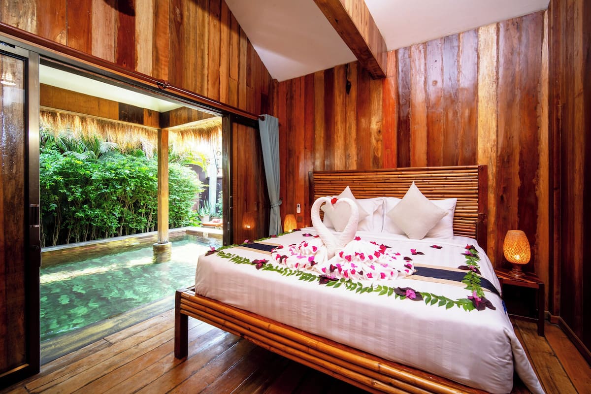 A wooden-paneled room with a bed, a nightstand, and a view of a lush garden through a glass door.