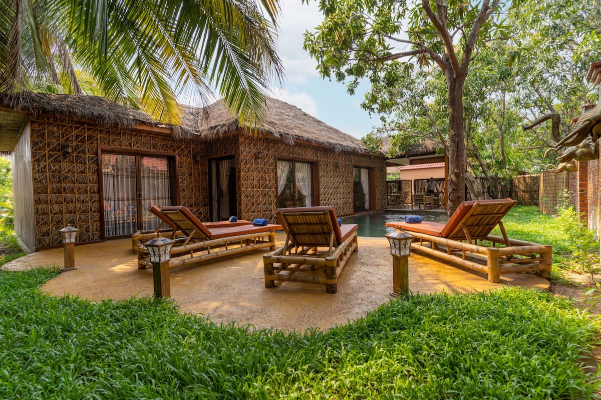 A thatched-roof bungalow with a pool, surrounded by lush greenery and bamboo furniture.