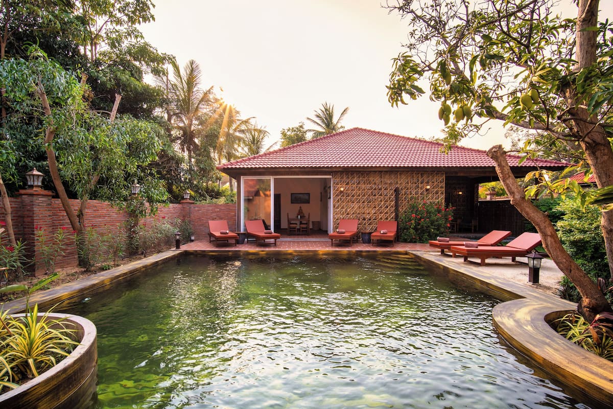 A pool with lounge chairs, a small building with a tiled roof, and surrounding greenery.