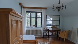 Ein Schlafzimmer mit Holzschrank, Bett mit weißer Bettdecke, Fenster mit Blick auf Bäume, Heizkörper, Tisch mit Stühlen und einer Pendelleuchte.