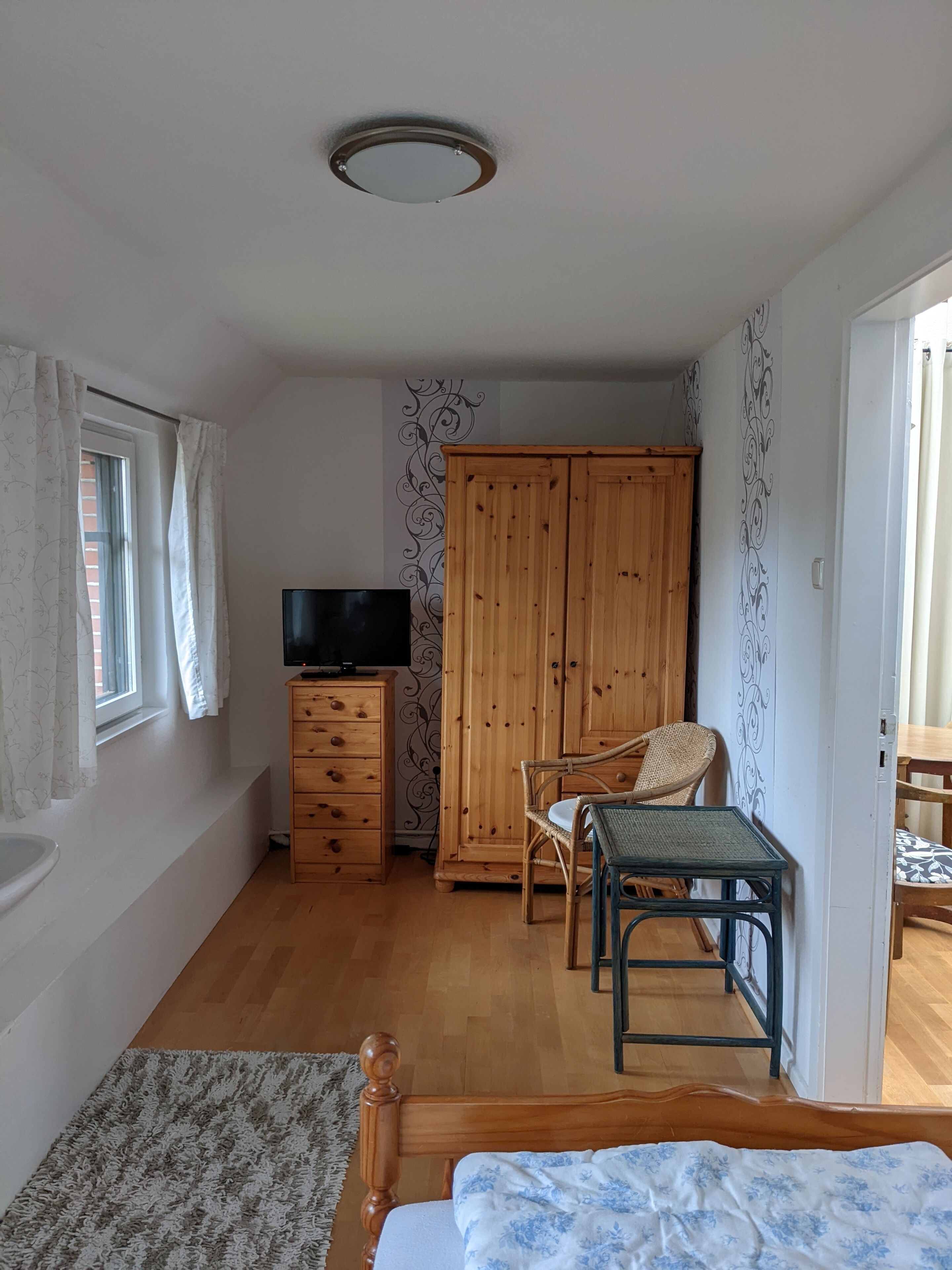 Een slaapkamer met een bed, een houten garderobe, een televisie op een dressoir, een raam met gordijnen en een kamer met een patroon op de muur.