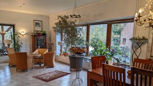 Ein gemütliches Wohnzimmer mit Korbmöbeln, einem Couchtisch und einem Kamin. Große Fenster mit Pflanzen bieten einen Blick ins Freie.
