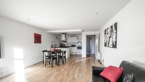 Een moderne woonkamer met een zwarte leren bank, een eethoek met een tafel en stoelen, en een keuken op de achtergrond.