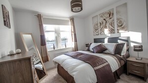 Een slaapkamer met een groot bed, een nachtkastje, een kaptafel, een raam met gordijnen en een aan de muur hangend kunstwerk.