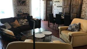 Una sala de estar acogedora con un sofá, un sillón y una mesa de centro de vidrio. Hay una zona de comedor con sillas y un mueble de madera.
