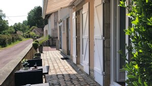 Un espacio de estar al aire libre con techo, mesas y sillas, un camino empedrado de piedra y un edificio con puertas y ventanas blancas.