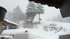 Un espace extérieur enneigé, avec un banc en bois et une table, entouré d’arbres chargés de neige et d’un bâtiment.