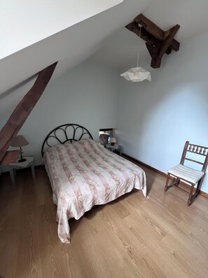 Un dormitorio con techo inclinado, una cama con colcha floral, una silla de madera, una mesita de noche con lámpara y una pequeña mesa.