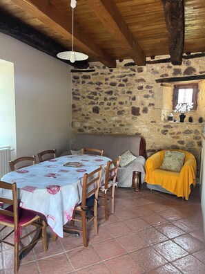 Un comedor rústico con pared de piedra, vigas de madera en el techo, una mesa redonda con sillas y un sillón amarillo con cojín.