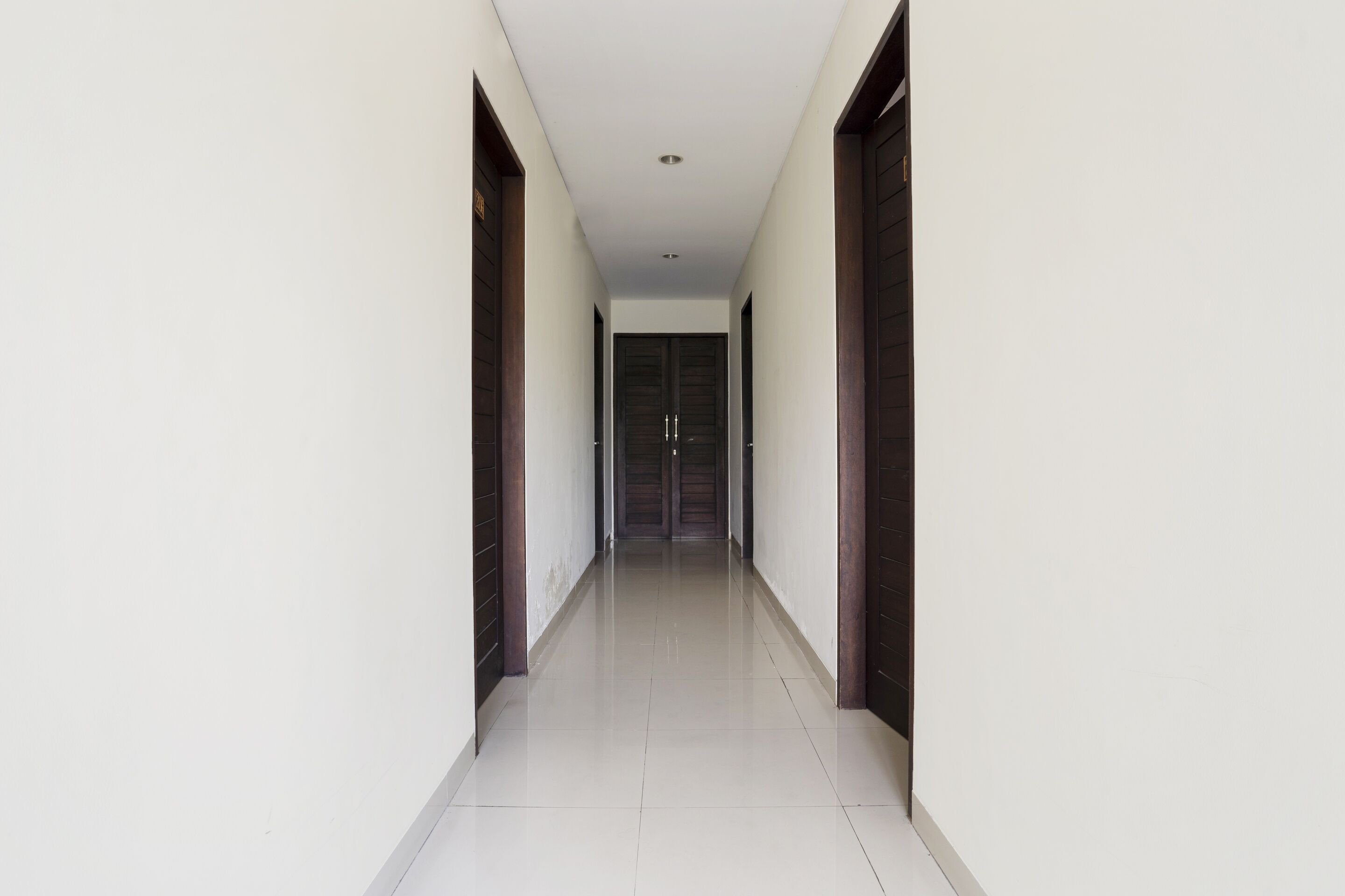 A long, narrow hallway with white walls and a shiny tiled floor, leading to a dark wooden door at the end.