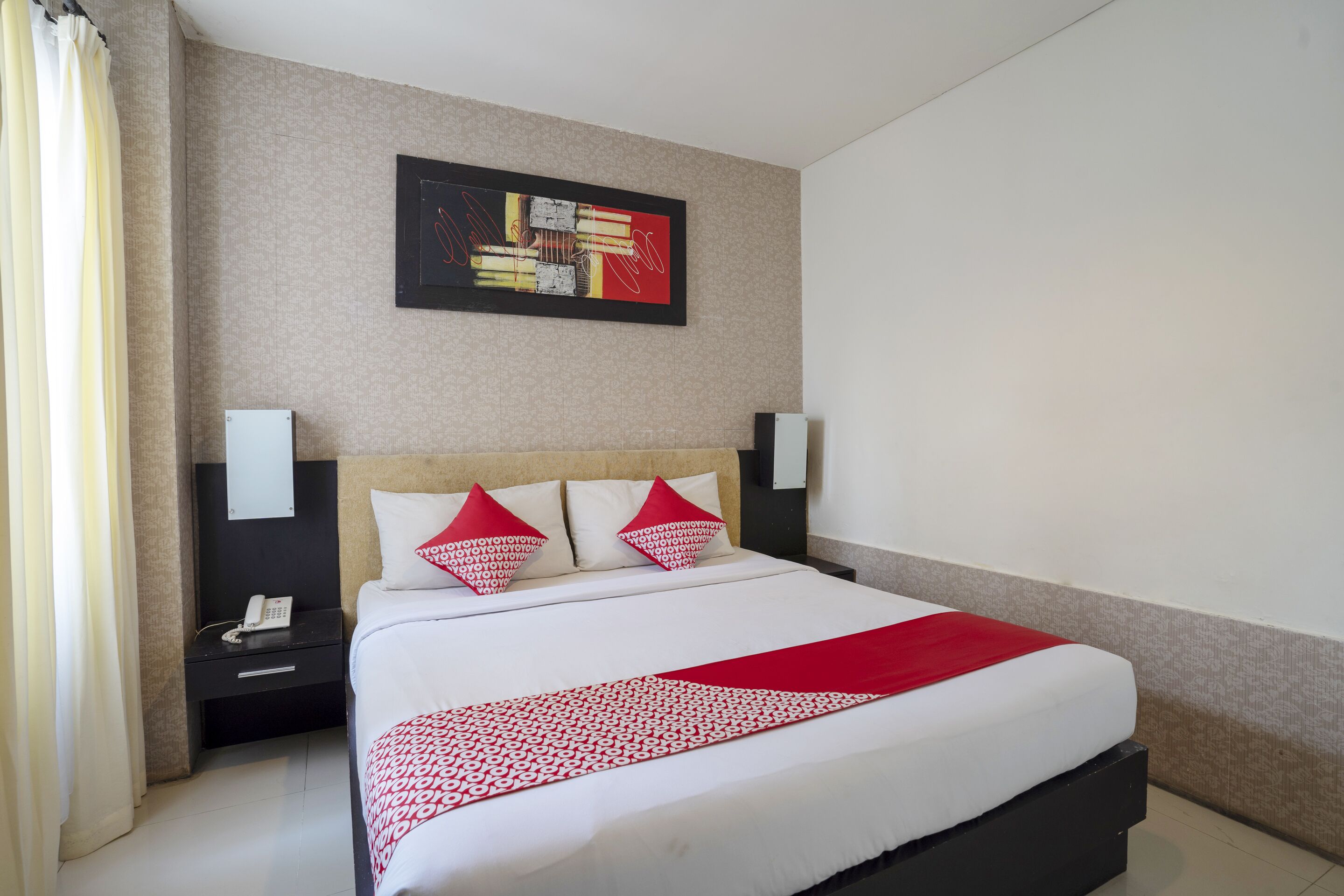 A hotel room with a bed, a nightstand with a phone, a wall-mounted artwork, and a red and white bedspread.