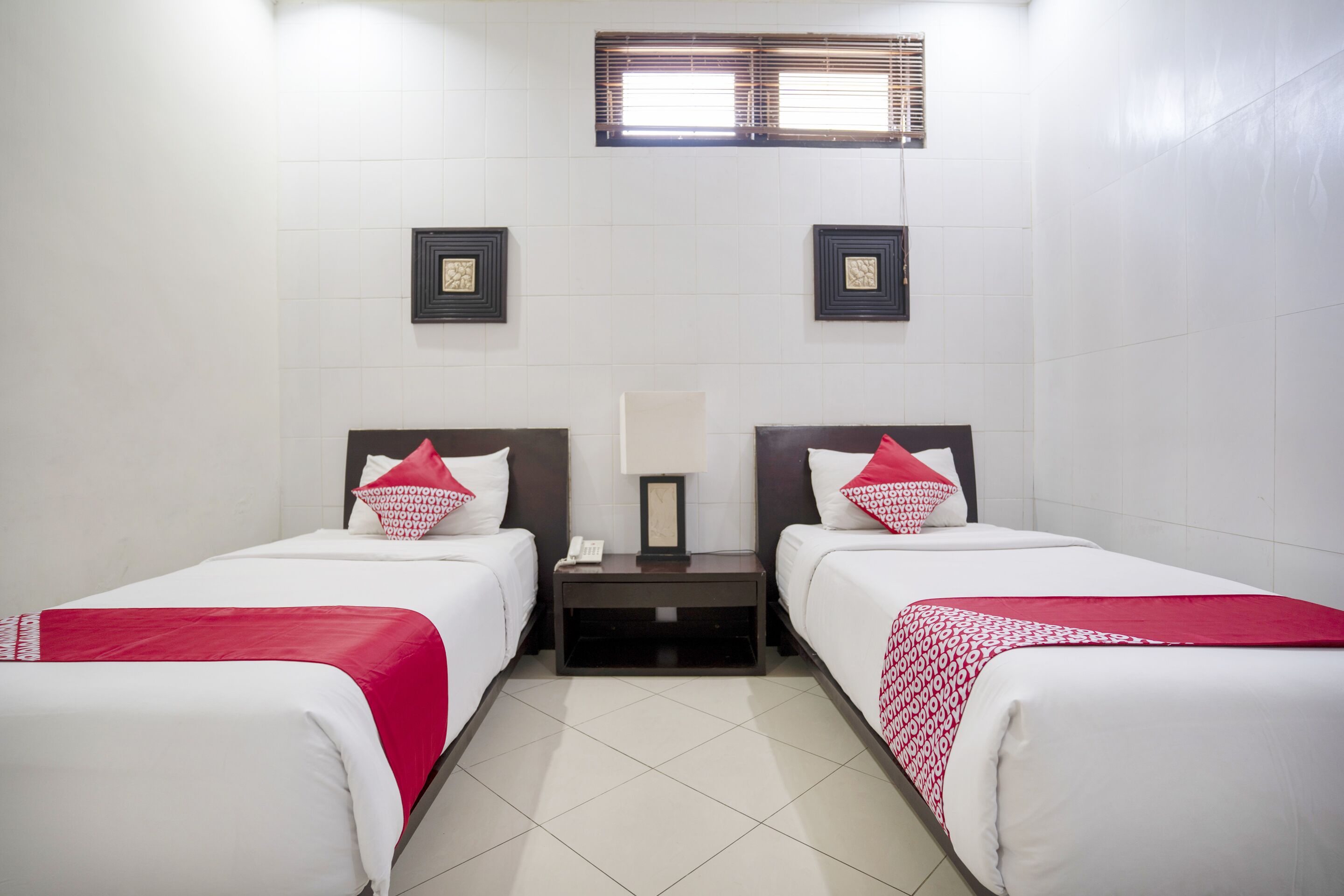 A hotel room with two beds, white bedding, red accents, a nightstand with a lamp, and framed pictures on the wall.