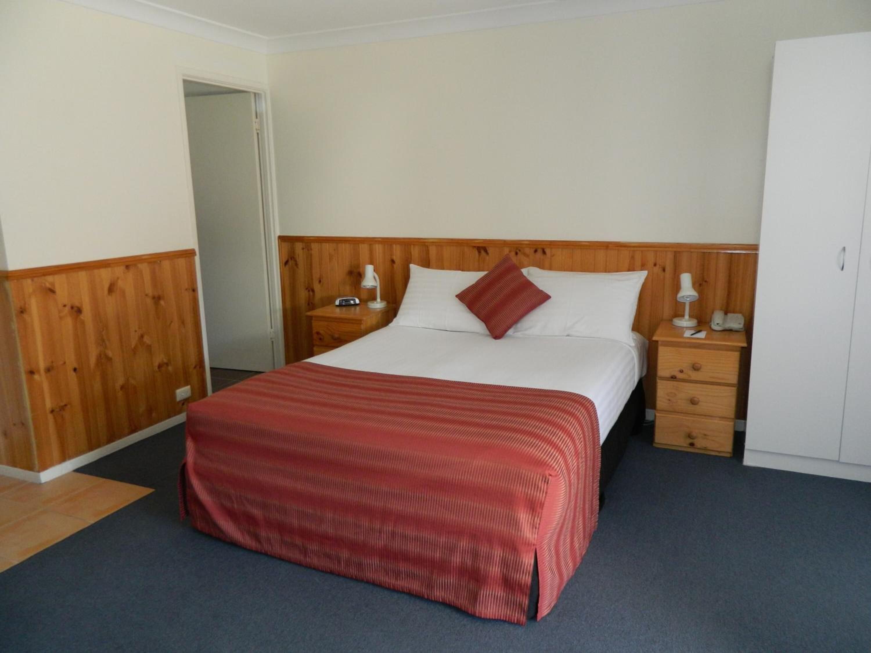 A hotel room with a wooden headboard, a single bed with a red blanket, two bedside tables with lamps, and a wardrobe.