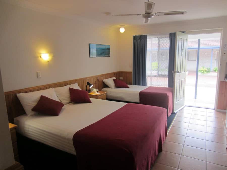 A hotel room with two beds, a ceiling fan, a nightstand with a lamp, and a view of the outside through a door.