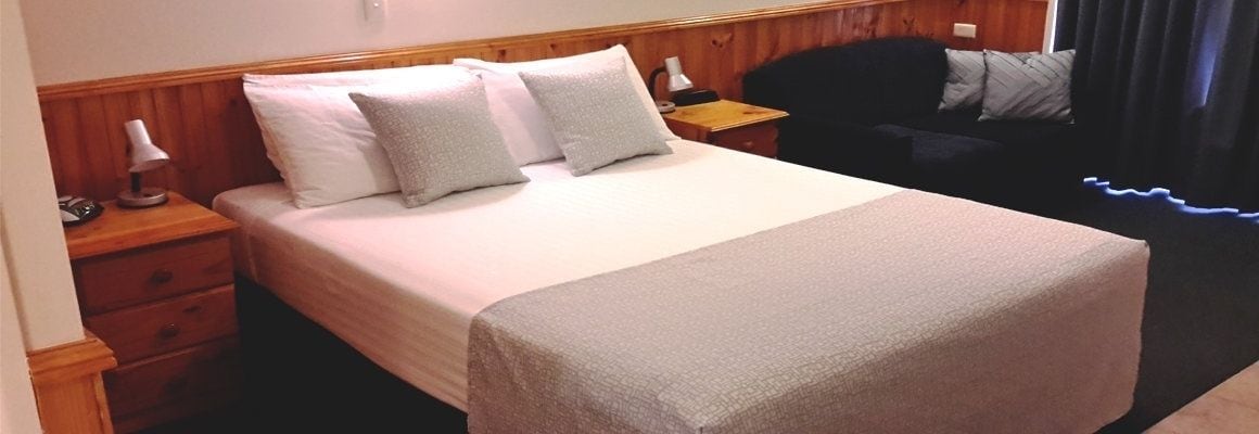 Una cama bien tendida con cubrecama blanco y gris, cabecera de madera, mesita de noche con lámpara y una silla negra en una habitación con paredes de madera.