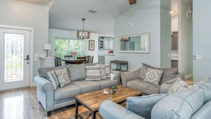 A spacious living room with a high ceiling fan, grey sofas, a wooden coffee table, and a view into the kitchen area.