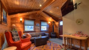 A cozy cabin interior with a wooden ceiling, a sofa, a chair, a table, and a fireplace.
