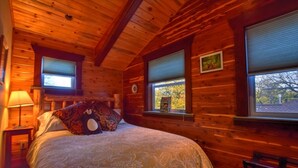 A wooden cabin bedroom with a bed, two windows with blinds, a nightstand, a lamp, and a chair.