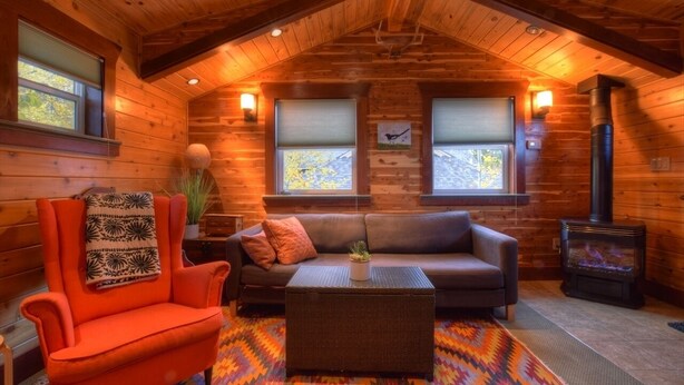 A cozy log cabin interior with a wooden ceiling, a sofa, an orange armchair, a fireplace, and two windows with blinds.