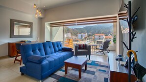 A modern living room with a blue sofa, a wooden coffee table, and a view of a cityscape through large windows.