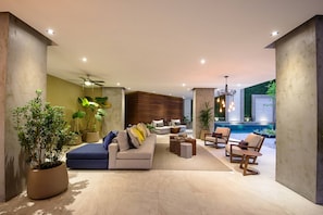 Un salon moderne avec un canapé, des fauteuils et une table basse. On y trouve une grande plante et une vue sur une piscine.