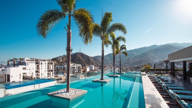 A rooftop infinity pool with palm trees, a clear view of the mountains, and modern buildings in the background.