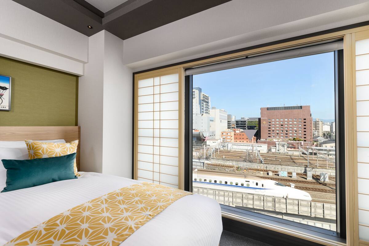Hotellihuone, jossa on suuri ikkuna, kuviollinen vuodevaatteilla varustettu sänky sekä näkymä kaupungin siluettiin ja rautatiekiskoille.