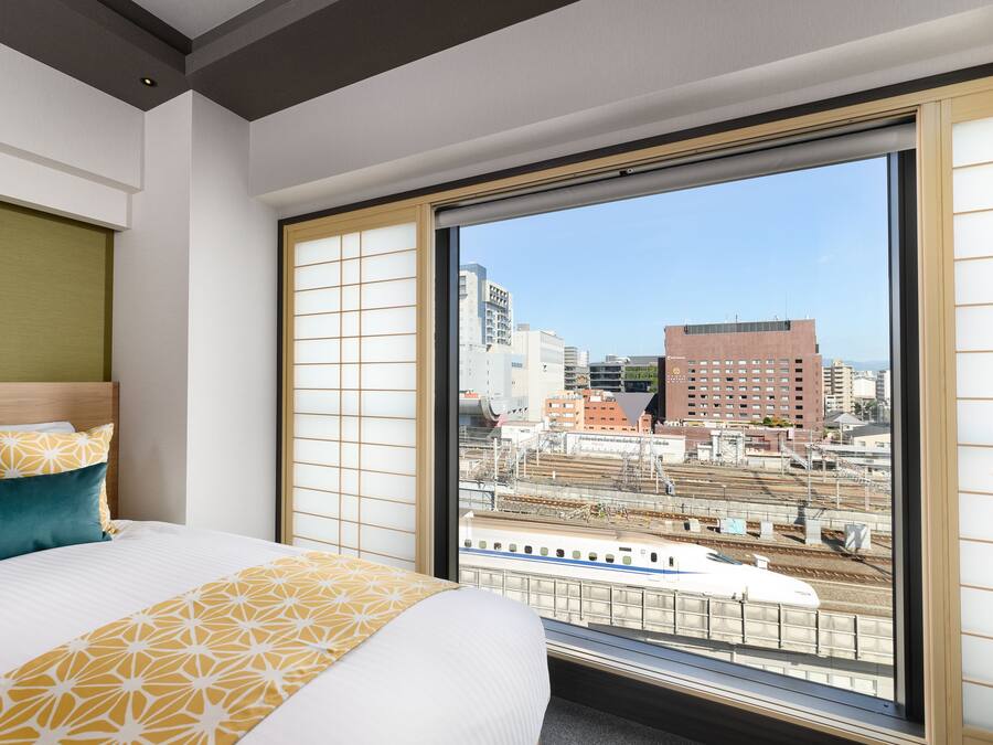 A hotel room with a large window, a bed with patterned bedding, and a view of the cityscape and train tracks.