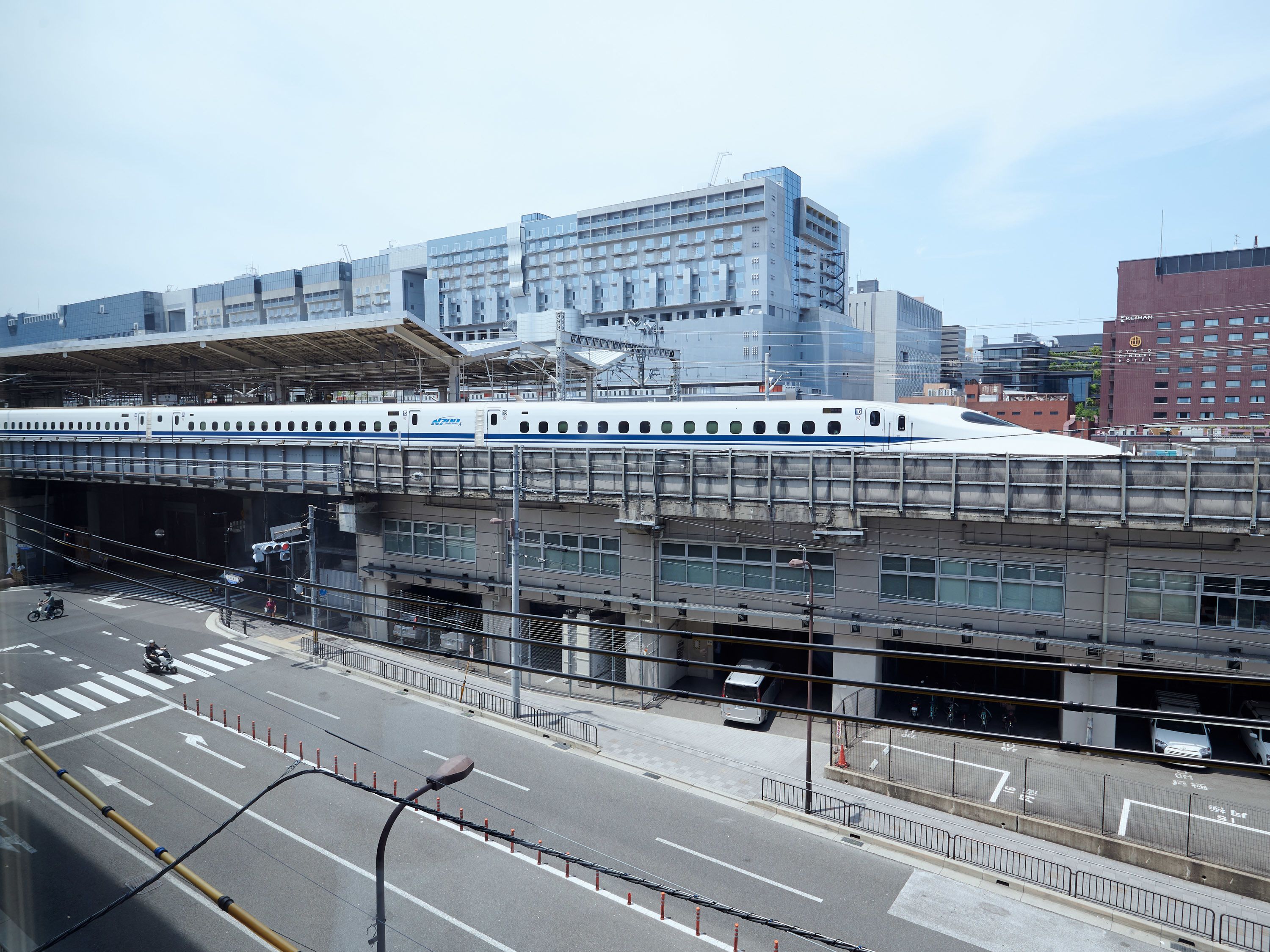 A train station with a modern train on the tracks, a pedestrian overpass, and various buildings in the background.
