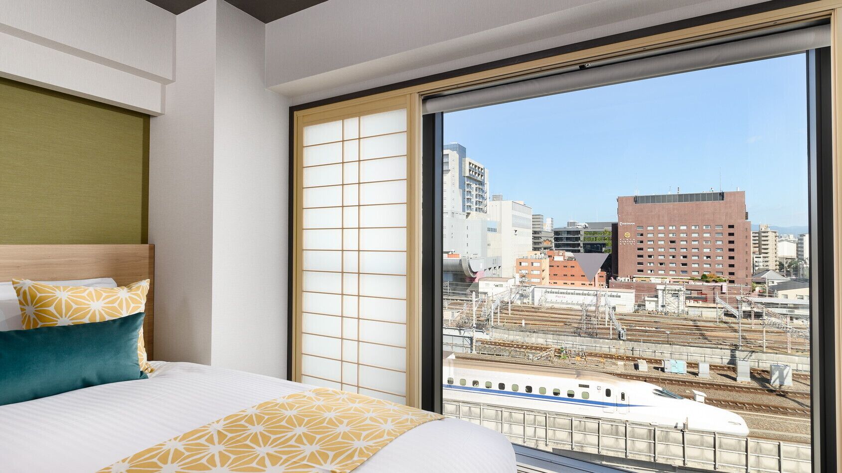 A hotel room with a large window, a bed with patterned bedding, and a view of the cityscape and train tracks.