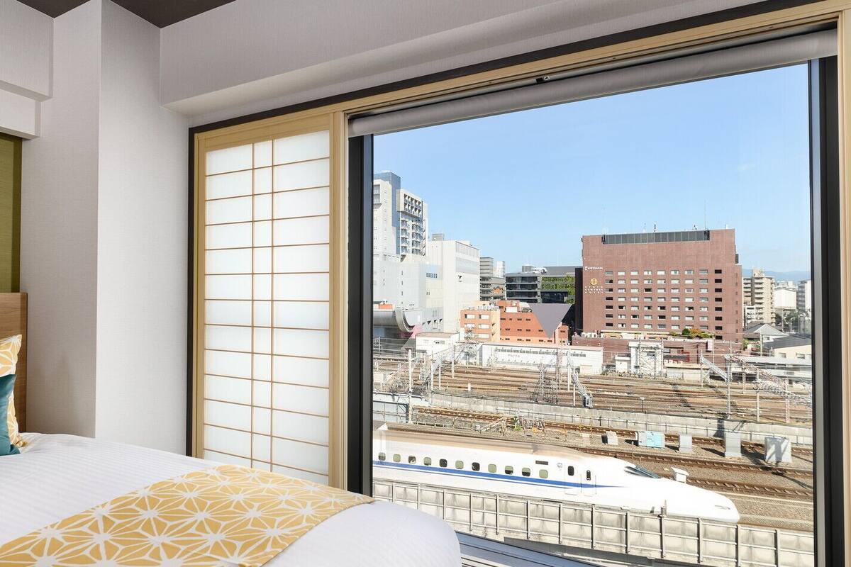 A hotel room with a large window, a bed with patterned bedding, and a view of the cityscape and train tracks.
