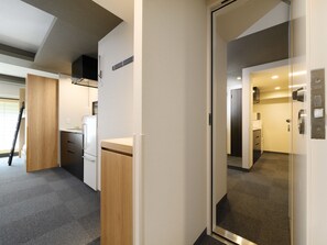 A modern hotel room with a kitchenette, a wooden cabinet, a mirror, and a door leading to another room.