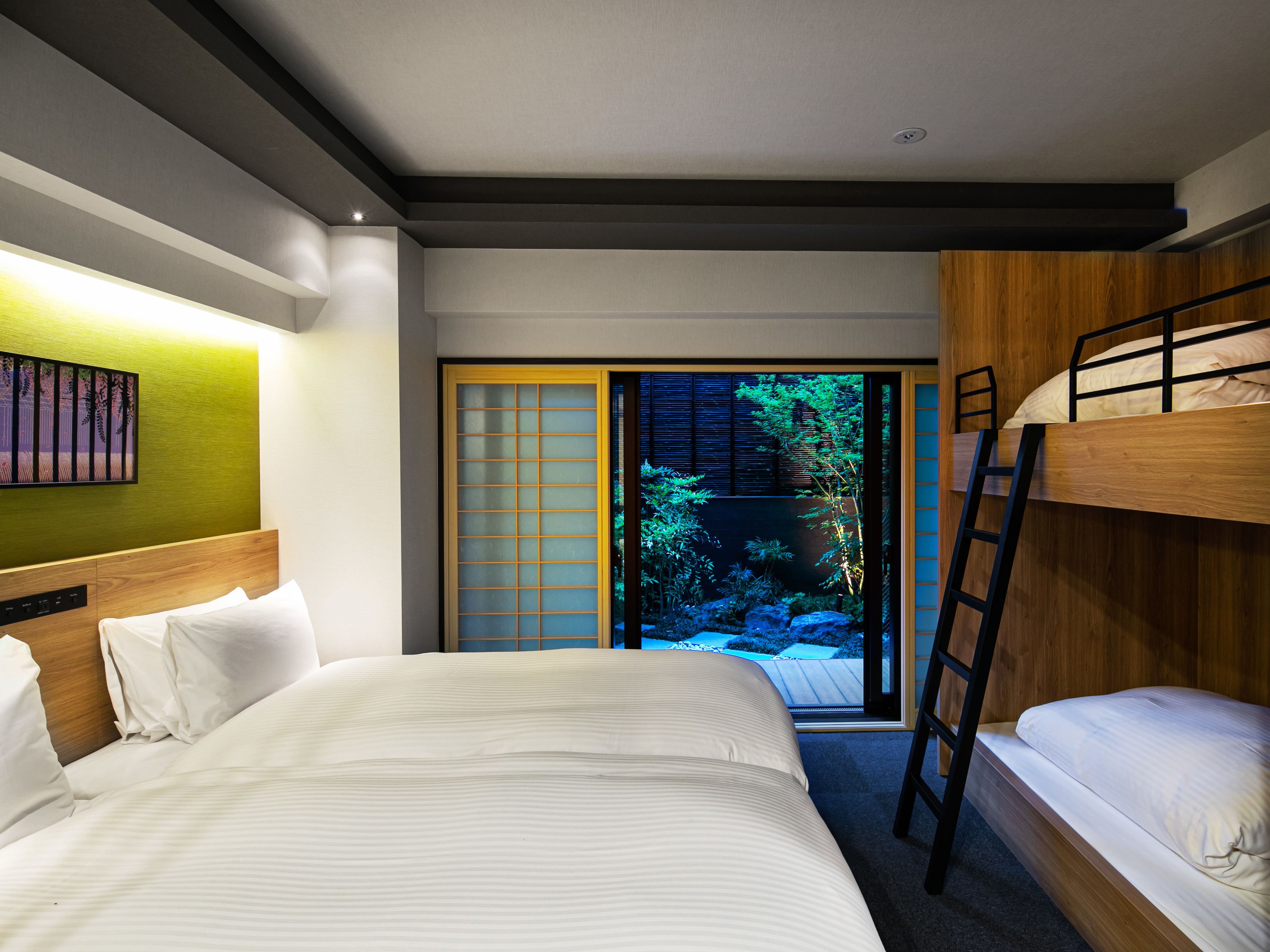 A hotel room with a large bed, a bunk bed, a ladder, a sliding door leading to a garden, and a wall-mounted artwork.