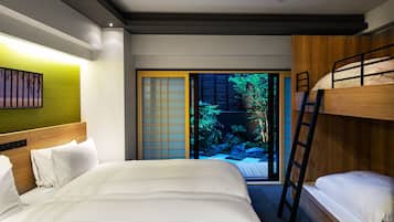 A hotel room with a large bed, a bunk bed, a ladder, a sliding door leading to a garden, and a wall-mounted artwork.