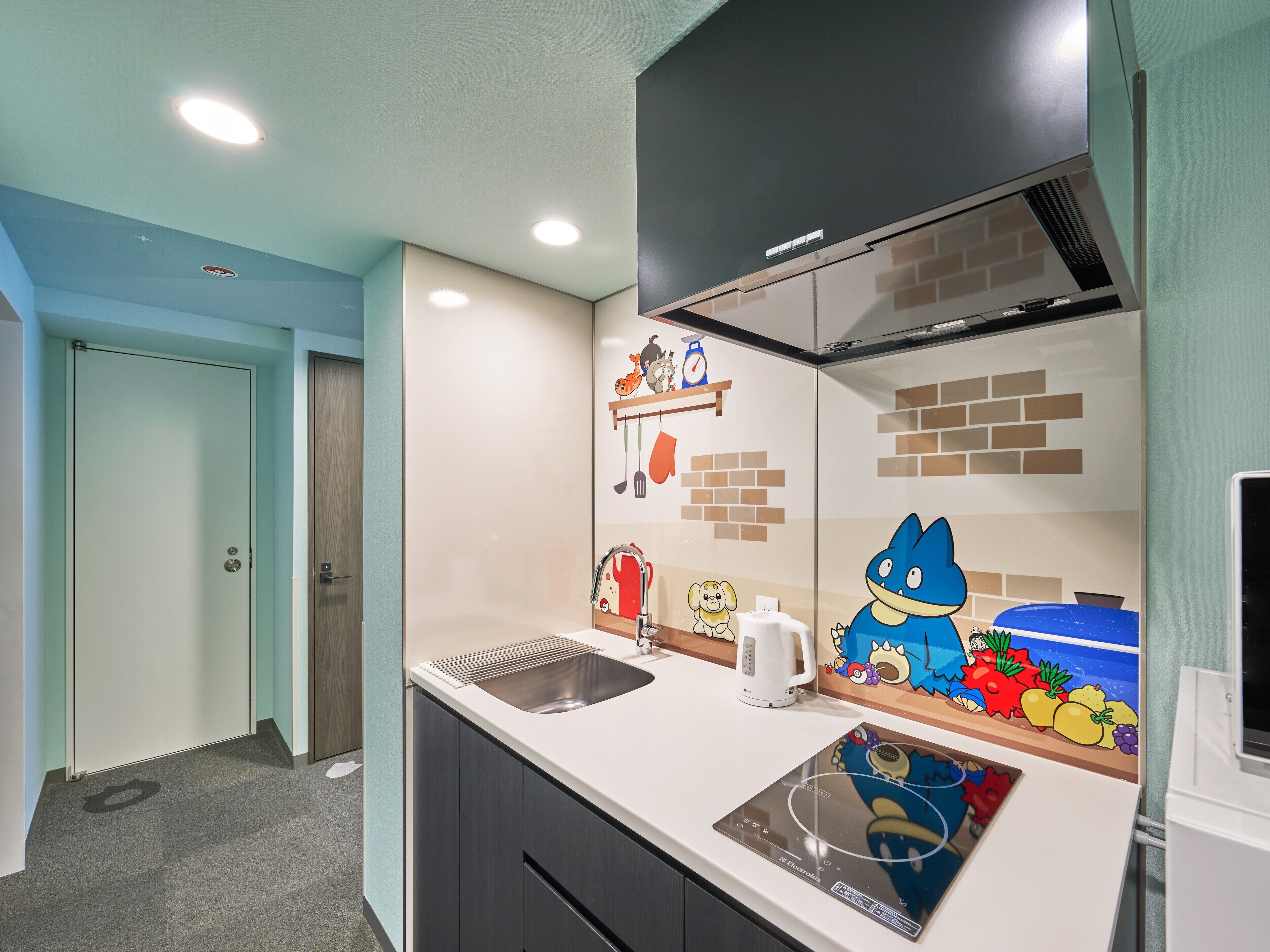 Una cocina moderna con estufa integrada, pileta y microondas. Se observa una pared decorativa con personajes de caricatura y una alacena con objetos de cocina.