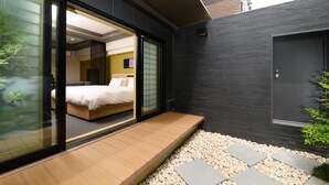 Une chambre d’hôtel moderne dotée d’une grande porte-fenêtre coulissante donnant sur un balcon, d’un lit avec du linge de lit blanc et d’un petit coin salon avec un banc en bois.