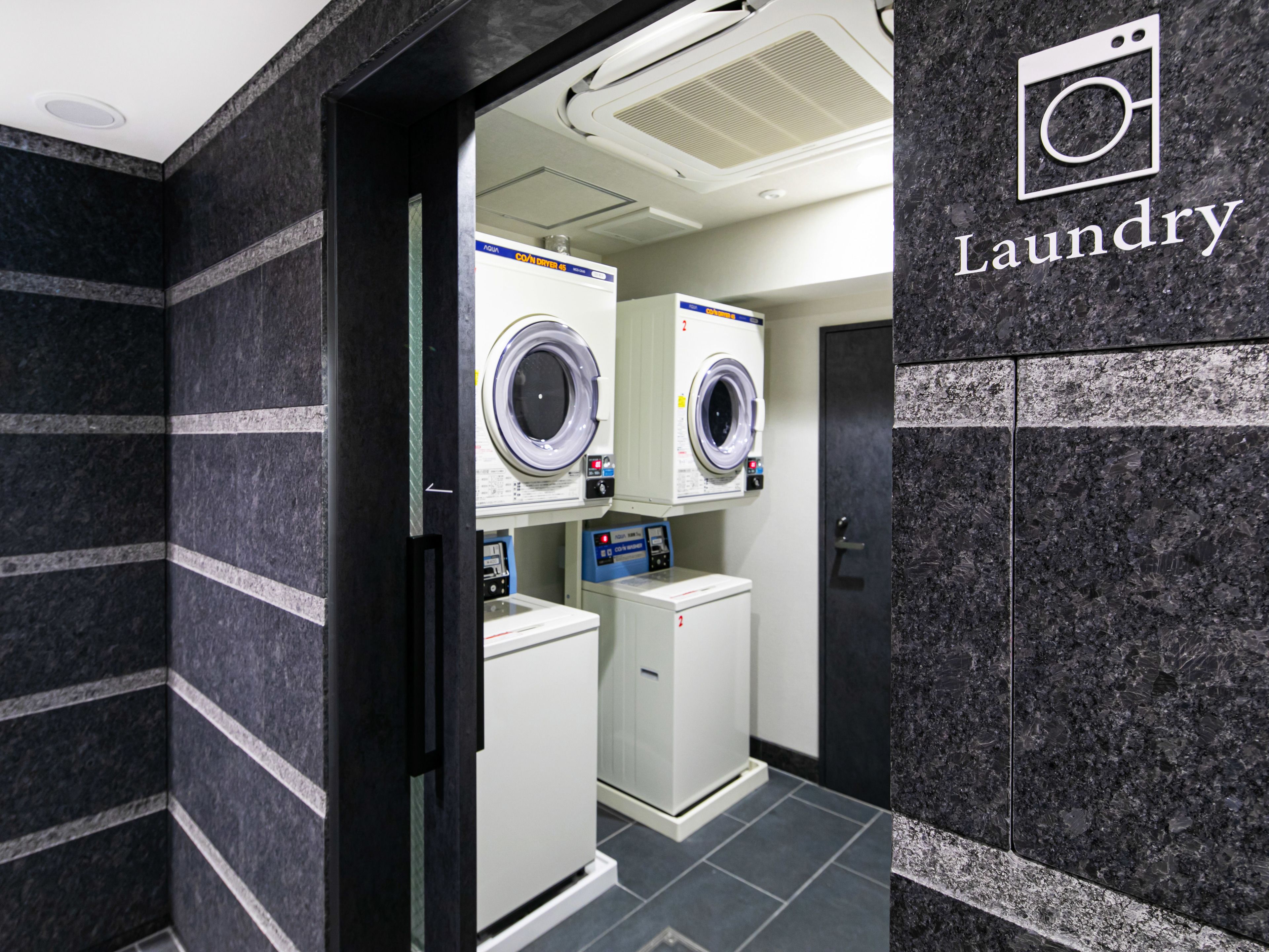 A laundry facility with two washing machines and a dryer, featuring a modern design with dark marble walls and a sign indicating the location.