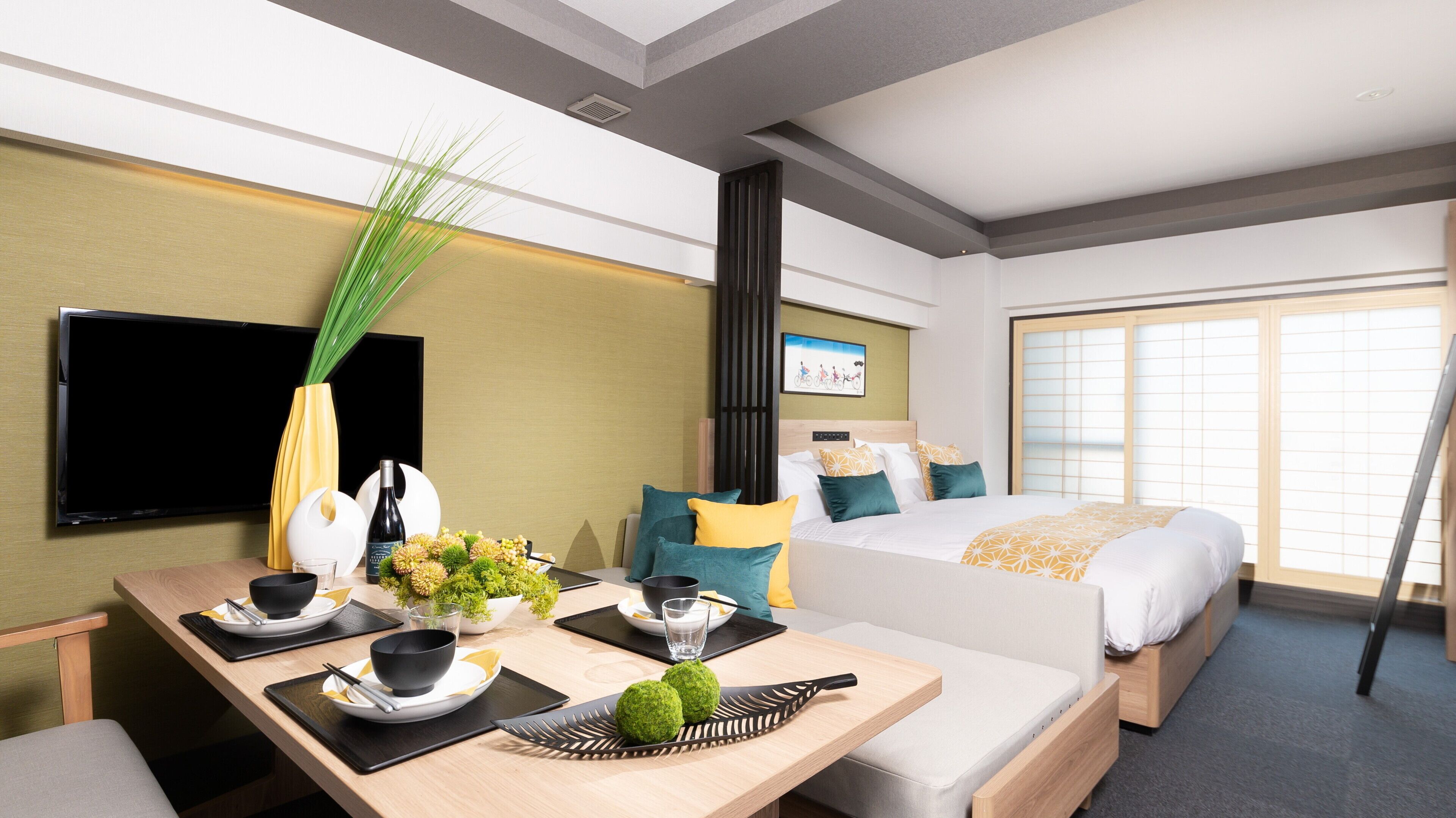 A hotel room with a bed, a TV, a dining area with a table set for two, and a large window.