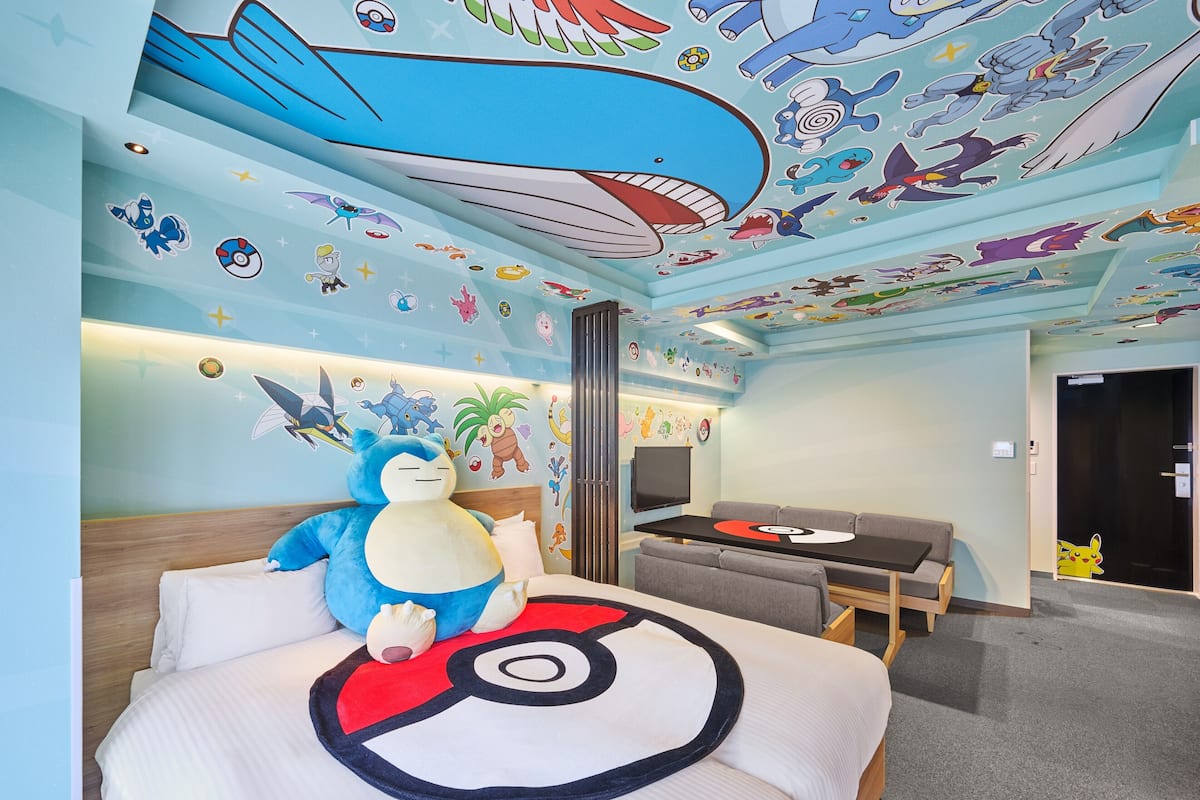 Hotellihuone, jossa on sänky, jonka päällä on Pokémon-aiheinen päiväpeite, suuri pehmolelu ja katossa oleva seinämaalaus, jossa on esitettynä erilaisia Pokémon-hahmoja.