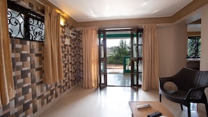 Habitación de hotel con suelo de baldosas, un sofá, una mesa con mandos a distancia y vistas a través de una puerta abierta.