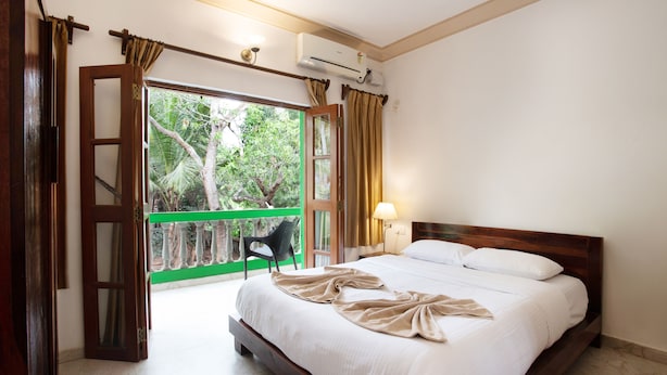 Una habitación de hotel con una cama grande, una puerta abierta que da a un balcón con vistas a árboles, y un equipo de aire acondicionado en la pared.