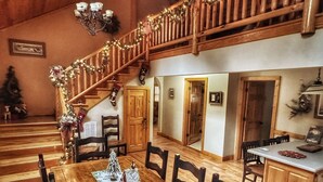 A wooden dining area with a long table, chairs, and festive decorations. A staircase with railings and lights leads to an upper level.