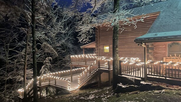 A wooden cabin with a covered deck and stairs, surrounded by snow-covered trees.