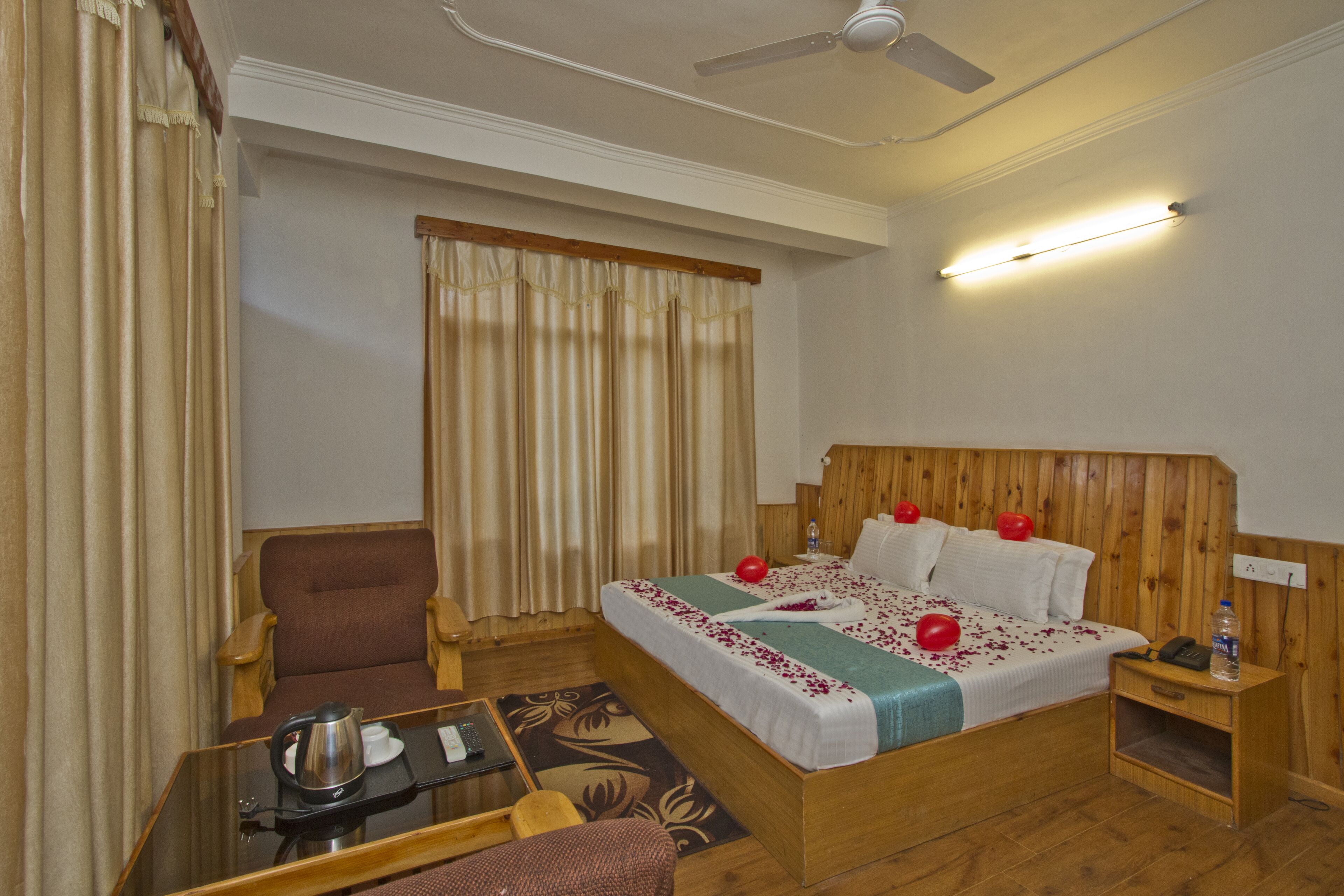 A hotel room with a wooden headboard, a bed with red pillows, a bedside table with a phone and a bottle, a brown armchair, a small table with a coffee pot and a cup, and a ceiling fan.
