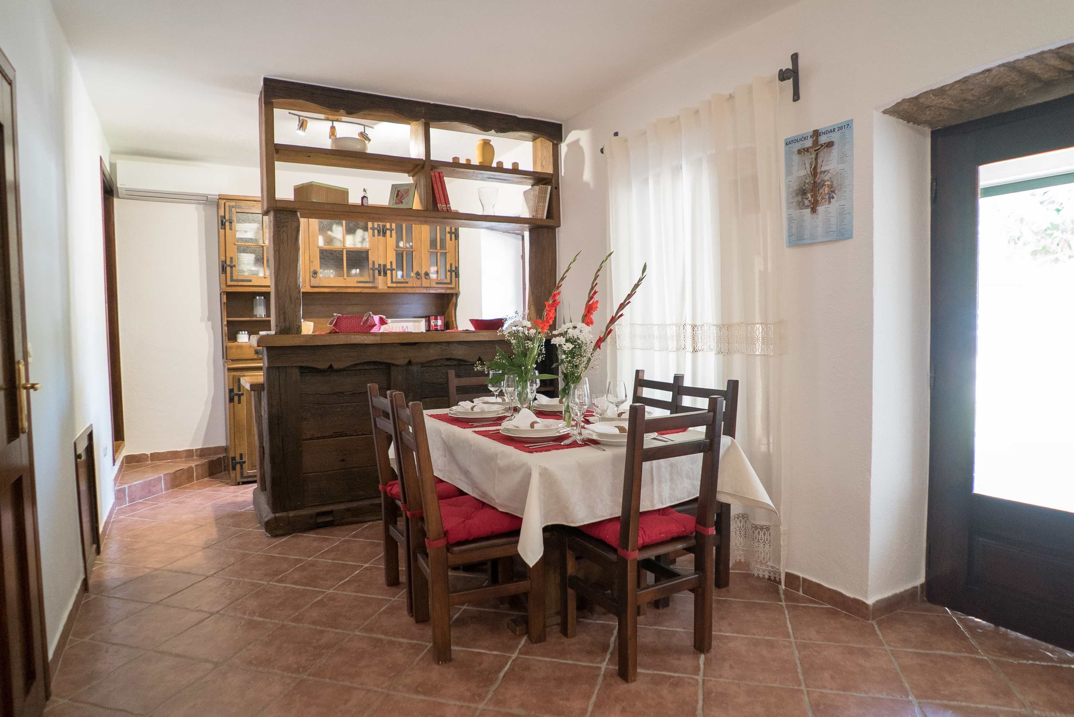 A dining area with a table set for four, surrounded by wooden chairs with red cushions. A wooden cabinet with glass doors is in the background, and a window with white curtains is visible.