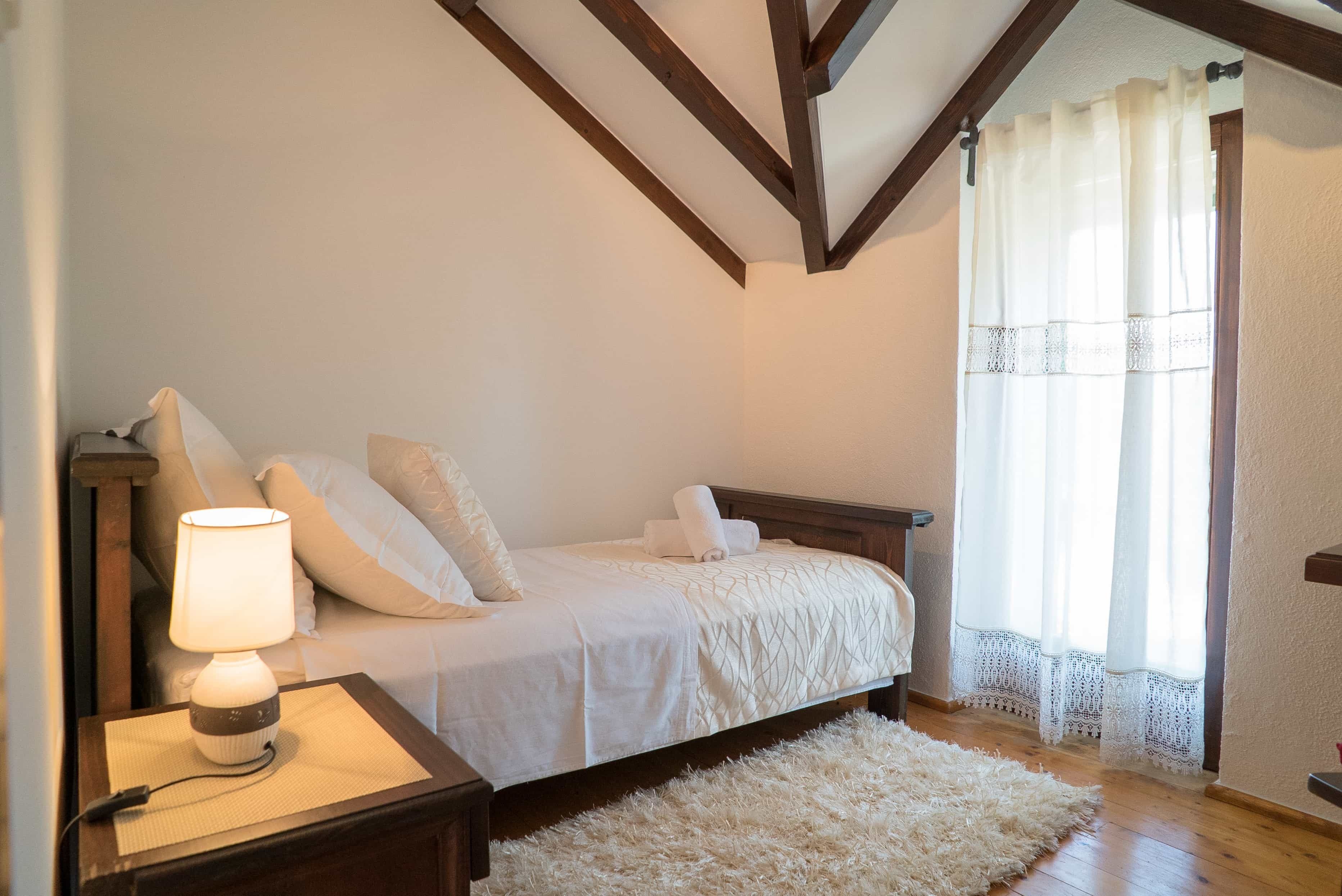 A bedroom with a bed, a nightstand with a lamp, a fluffy rug, wooden beams on the ceiling, and a window with sheer curtains.