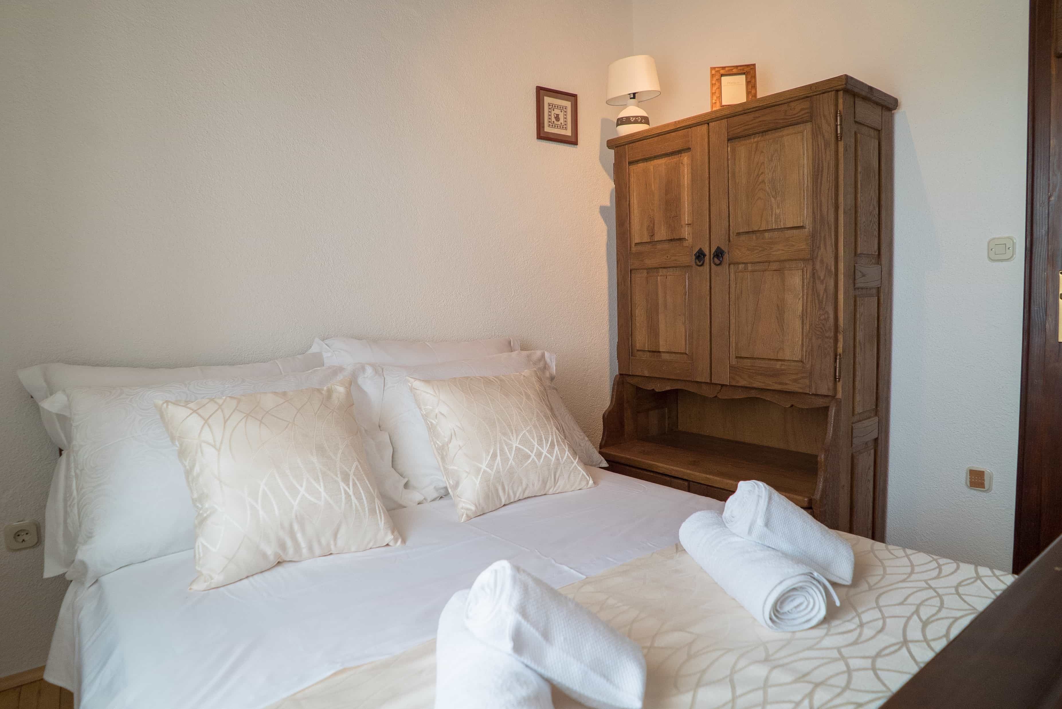 A neatly made bed with white linens and pillows, a wooden wardrobe, and a small table with a lamp and picture frame.