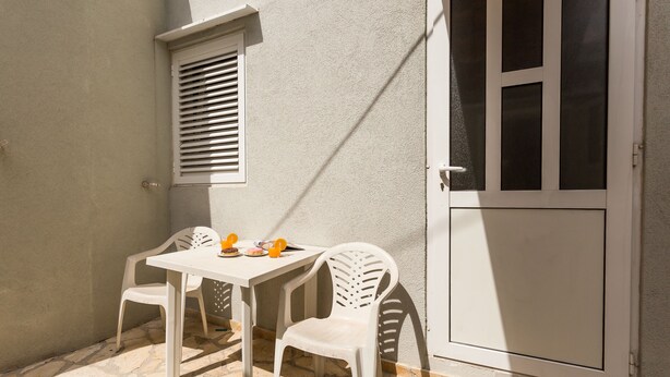 Une petite terrasse extérieure avec une table et deux chaises, une fenêtre avec des stores et une porte.