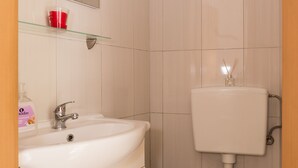 Une salle de bain avec un lavabo, un miroir et des toilettes blancs. On y trouve un panier bleu et une tasse rouge posés sur le lavabo.