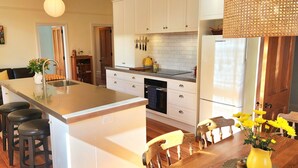 A modern kitchen with white cabinets, a central island with stools, and a dining area with wooden chairs and a yellow vase. - Rewa House ~ city home, 3 bedrooms, walk to CBD (Christchurch)